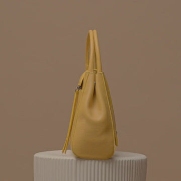 A fashion video showing a rotating yellow leather handbag. The handbag has a classic trapeze shape with a straight quilted flap in the middle and top handles. This is the front view of the Tomoli Briffani Lean interchangeable tote handbag in Maize. This bag can be used as an everyday bag, a work tote, or even an oversized clutch.
