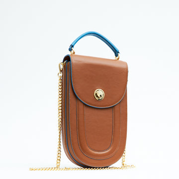 A fashion product photo showing the front view of a colorful brown leather handbag. The bag has an elongated saddle shape with a flap closure, contrasting blue trims, and a blue top handle. There is a chain strap and a  metal lock in gold hardware. This is the Tomoli Fitini II structured saddle handbag in Sky Pecan. This handbag can be used as an everyday crossbody bag, a work bag, or a special occasion bag.