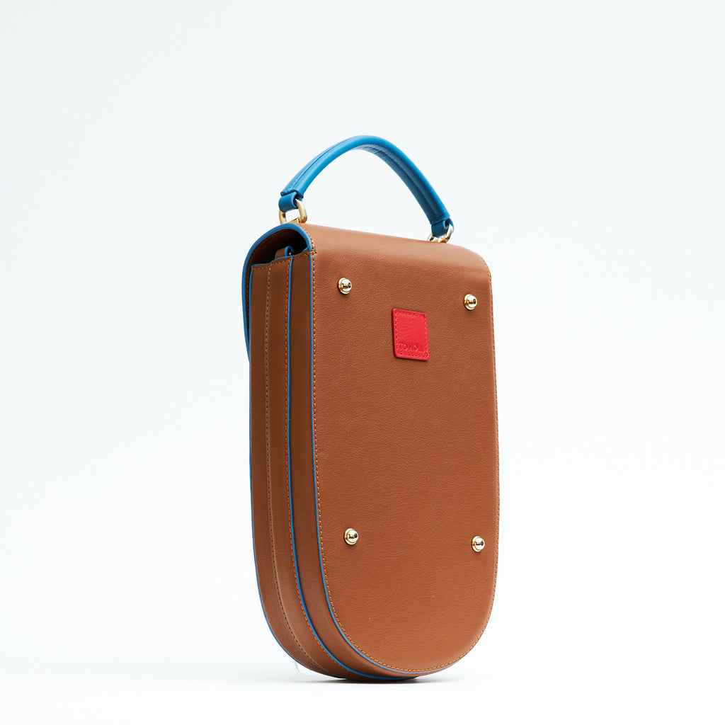 A fashion product photo showing the back view of a colorful brown leather handbag. The bag has an elongated saddle shape with blue trims and a blue top handle. There is a small red square and gold metal rivets. This is the Tomoli Fitini II structured saddle handbag in Sky Pecan. This handbag can be used as an everyday crossbody bag, a work bag, or a special occasion bag.
