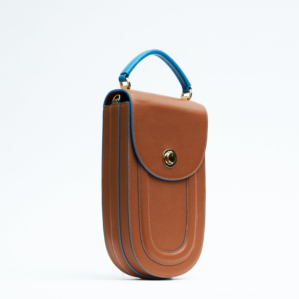 A fashion product photo showing the side view of a colorful brown leather handbag. The bag has an elongated saddle shape with a flap closure, contrasting blue trims, and a blue top handle. There is a metal lock in gold hardware. This is the Tomoli Fitini II structured saddle handbag in Sky Pecan. This handbag can be used as an everyday crossbody bag, a work bag, or a special occasion bag.